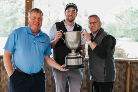 Three men, including one in a "BOWERS" shirt, pose with a large silver golf trophy in a wooden pavilion.