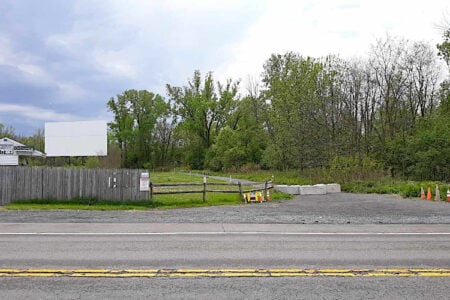 A drive-in movie theater's screen and entrance booth stand behind a fence, bordering a rural road and woods.