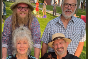 Four smiling individuals, three men and one woman, pose outdoors on a grassy lawn with large brick buildings behind them.