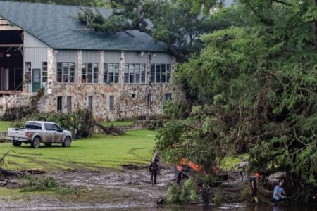 Crews clear fallen trees and debris from muddy ground near a storm-damaged building and pickup truck.