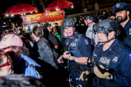 NYPD tactical officers with batons stand among a dense nighttime crowd near a food truck.