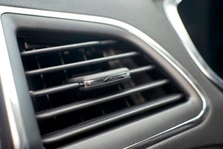 Close-up of a car's black air vent with chrome trim, featuring a small dark air freshener clipped to a louver.