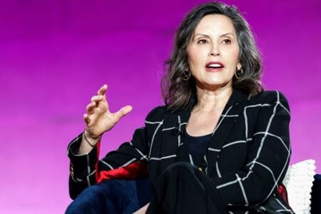 Gretchen Whitmer, Michigan Governor, speaks and gestures, seated in a blue chair against a bright purple background.