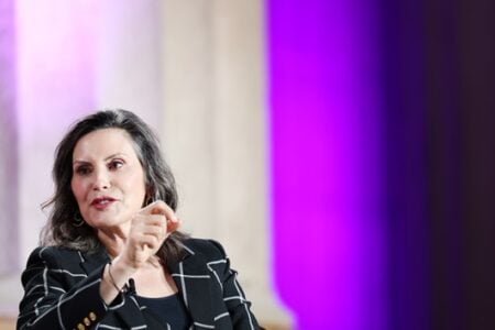 Gov. Gretchen Whitmer speaks, gesturing with her right hand, against a backdrop of glowing purple lights and columns.