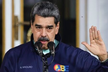 Venezuelan President Nicolas Maduro speaks at a podium, raising his right hand with a fly visible, indoors.
