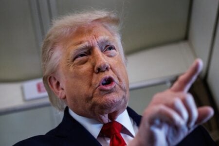 Donald Trump, in a suit and red tie, speaks with open mouth, pointing his right index finger in an airplane cabin.
