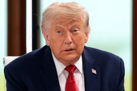 Donald Trump, wearing a dark suit, red tie, and flag pin, looks forward with his mouth slightly open, indoors.
