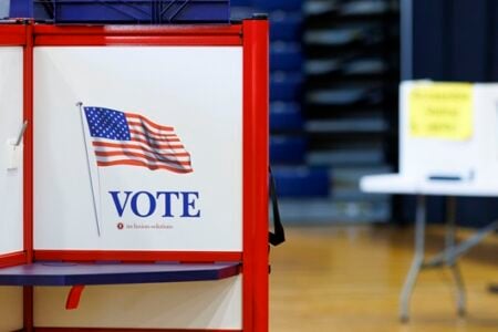 A red and white voting booth displaying an American flag and "VOTE" sign in a gymnasium polling place.