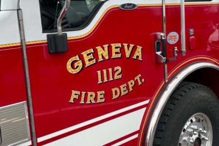 Side of a red Geneva Fire Department truck, featuring gold lettering 'GENEVA 1112 FIRE DEPT.' and chrome accents.