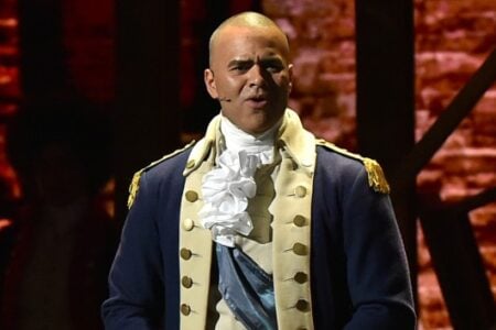 Shaved-head actor in blue Revolutionary War uniform with ruffled cravat, singing into a microphone on stage.