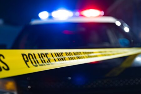 Yellow police line tape foregrounds a police car with flashing red and blue lights at a nighttime incident.
