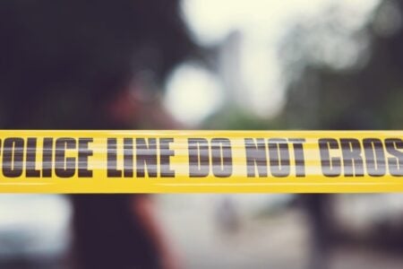 Yellow "POLICE LINE DO NOT CROSS" tape blocks the foreground of a blurred outdoor scene.