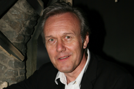 A man with graying hair and an earring smiles at the camera in a dimly lit room with a stone wall.