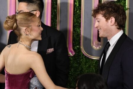 Ariana Grande in a magenta dress and Ethan Slater in a suit smile at each other against a green 'Wicked' premiere backdrop.