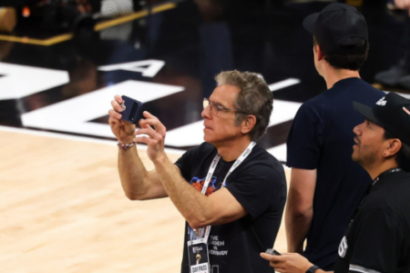 Ben Stiller, wearing glasses, holds a phone to record from courtside during the NBA Finals.