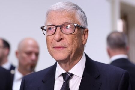 Close-up of Bill Gates, wearing glasses and a dark suit, looking slightly left at an indoor event.