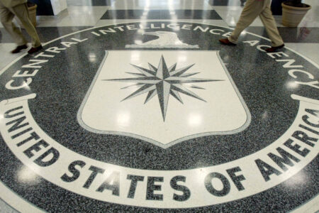 Two people's legs stride across the Central Intelligence Agency seal embedded in a polished dark and light terrazzo floor.