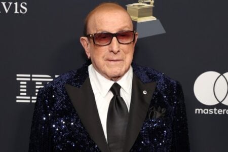 Music executive Clive Davis poses in a blue sequined blazer and sunglasses before sponsor logos and a Grammy award.