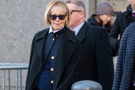 Woman with blonde hair, sunglasses, and a navy coat with gold buttons over a white collar walks past a building.