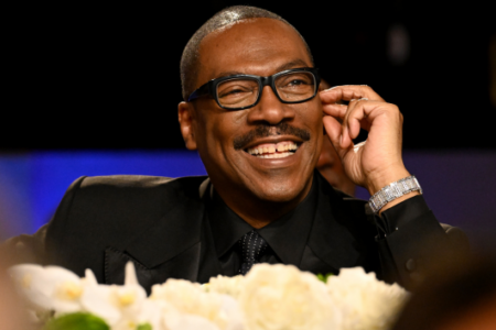 Comedian Eddie Murphy, in a suit and glasses, smiles widely at an indoor event.