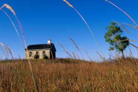 Small stone building with a red-capped cupola on a hill covered in tall dry grass, with a tree under a blue sky.