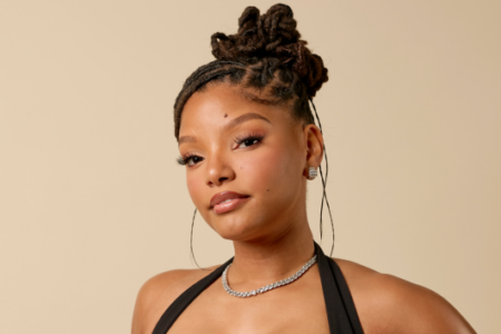 Halle Bailey, with locs styled in an updo, wears a diamond necklace and black top while posing against a tan background.