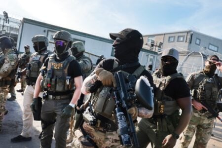 Masked tactical ICE & police officers with less-lethal weapons stand guard at a fenced facility.
