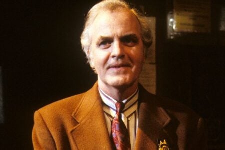 White-haired man in brown jacket, striped shirt, and patterned tie smiles, wearing a medal against a dark background.