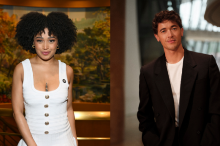 A woman with curly hair in white smiles before a painting, next to a man in a black blazer in a modern building.