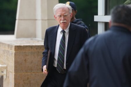 John Bolton, former National Security Advisor, walks past a stone wall and building entrance in a suit.