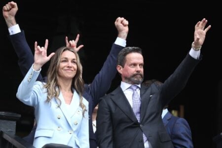 Smiling woman in a light blue blazer makes a "love you" sign next to a man in a dark suit waving from a balcony.