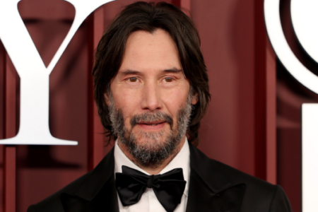 Bearded Keanu Reeves looks at the camera with a slight smile, wearing a tuxedo and bow tie against a red event backdrop.