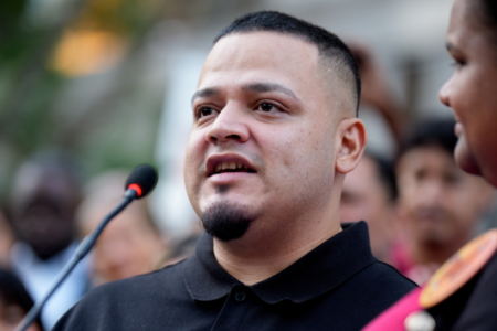 A man in a black polo shirt, with a fade haircut and goatee, speaks into a microphone at an outdoor event.