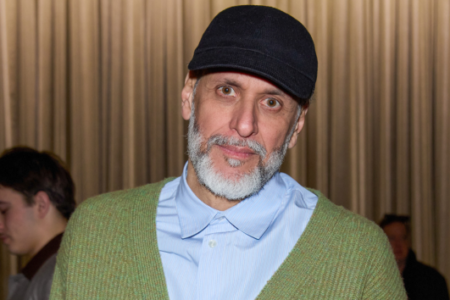 Man with grey beard, black cap, green cardigan, and blue shirt looks intently in front of beige curtains.