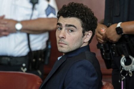 Man in dark suit with curly hair looks toward camera from court bench, flanked by officers.