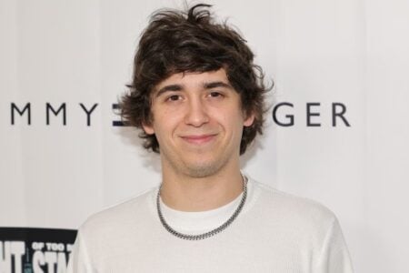Young man with messy brown hair and silver chain smiles against a white Tommy Hilfiger logo backdrop.