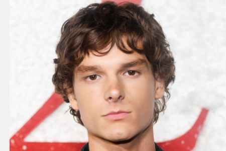 Young man with curly brown hair looks directly at the camera, against a white textured background with red lettering.
