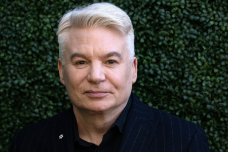 Mike Myers with light blonde hair and a dark pinstripe suit smiles against a green leafy backdrop.