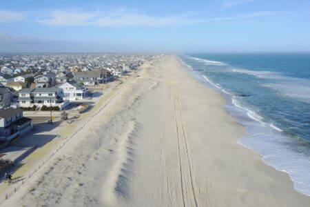 Aerial view of a wide sandy beach, ocean waves, a dense coastal town, and two people biking on a path.