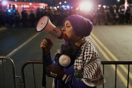 Woman in purple head covering and keffiyeh speaks into a megaphone on a street at night, facing police lights.
