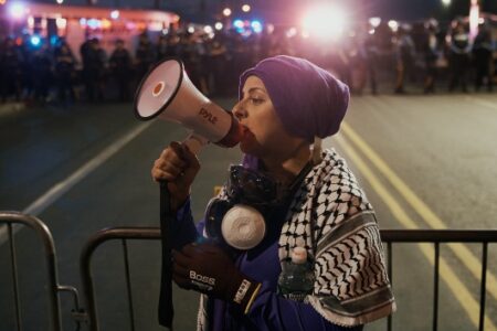 Woman in purple headscarf, keffiyeh, and respirator speaks into a megaphone at night, facing police.