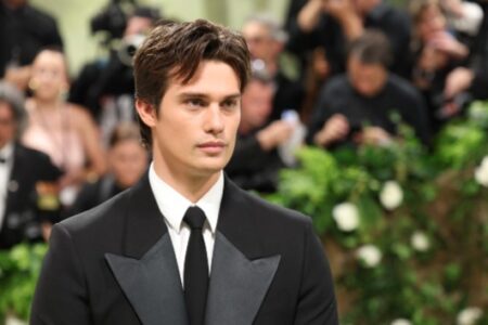 A young man in a black tuxedo stands with a focused expression at a crowded formal event with lush greenery.