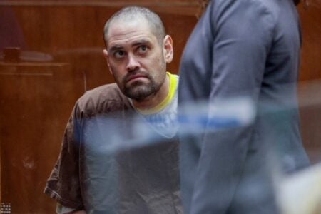 Bearded man in brown shirt looks up from behind glass in a courtroom; another person partially visible.