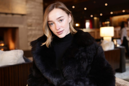 Young woman in a black fur coat and turtleneck smiles at the camera, seated in a warm lounge with a fireplace.