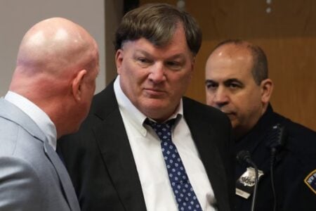 A man in a suit and patterned tie looks toward another man in a courtroom, with a NY State Courts officer present.