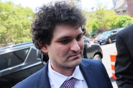Sam Bankman-Fried, wearing a suit and striped tie, looks down outdoors on a street with cars and trees.