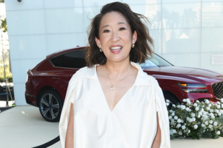 Sandra Oh smiles in a cream top, standing outdoors next to a red SUV decorated with white flowers at an event.