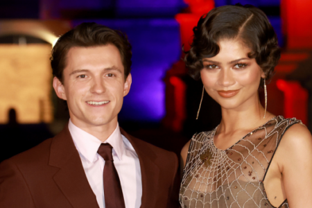 Actors Tom Holland in a brown suit and Zendaya in a spiderweb dress smile at an evening event.