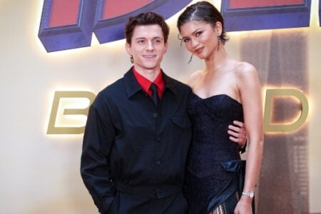 Tom Holland and Zendaya stand smiling side-by-side at an event, with illuminated "HOME" letters in the background.