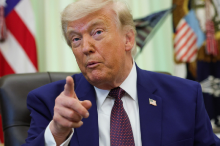 Donald Trump points emphatically with his right hand while seated in the Oval Office, wearing a suit and tie.